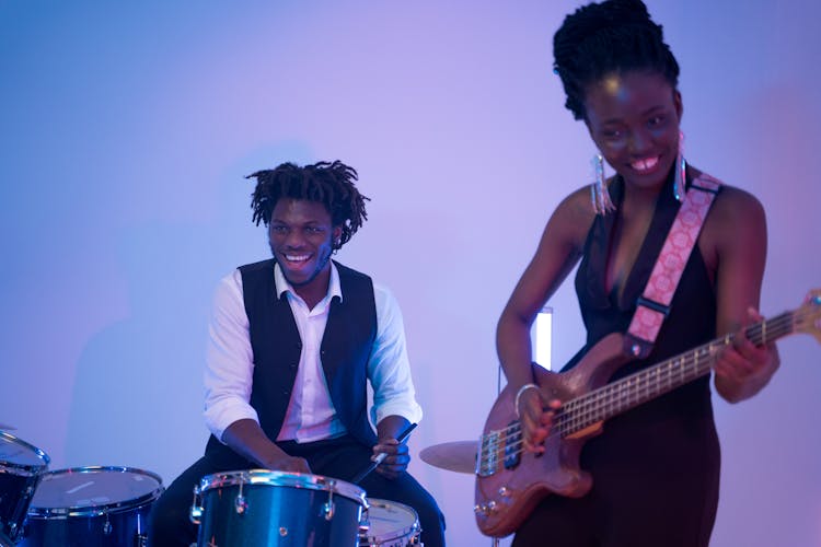 Man In White Button Up Shirt Playing Drums Beside A Woman Playing Electric Guitar