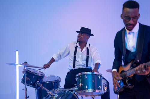 Man in White Dress Shirt Playing Drums Beside a Man Playing Electric Guitar