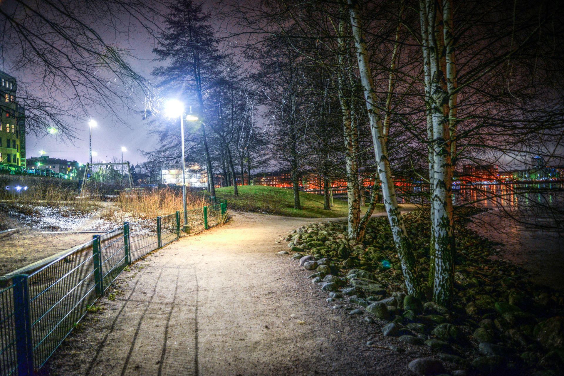 Illuminated park path at night, lined with birch trees and a distant cityscape reflection on the river.