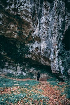 A lone traveler stands at the entrance of a rocky cave surrounded by autumn foliage.