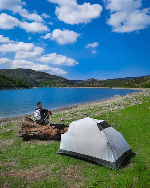 A Person Sitting on a Wooden Log Beside a White Tent Near a Lake