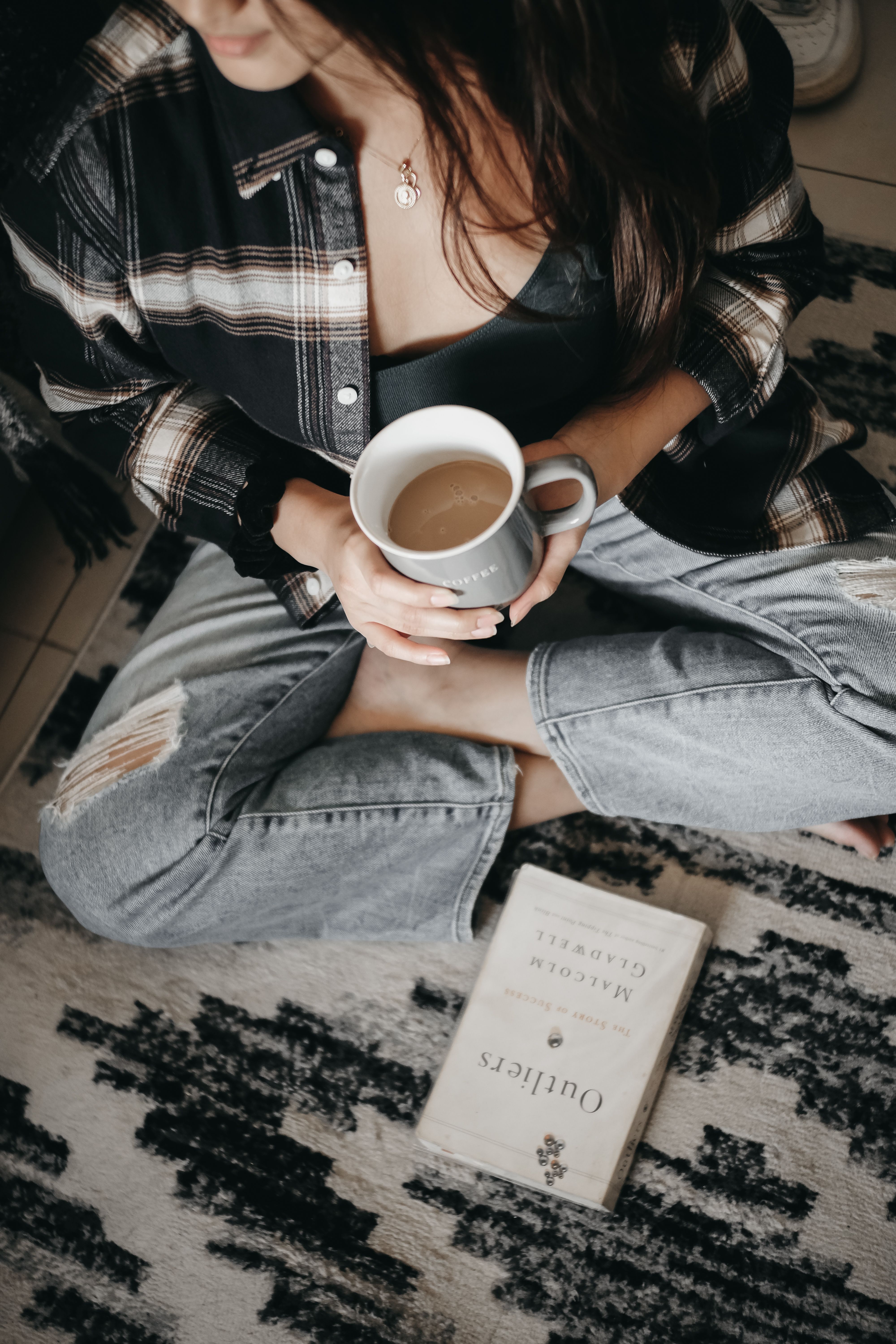 Woman sitting cross-legged with coffee and book, enjoying a calm morning on a patterned rug.