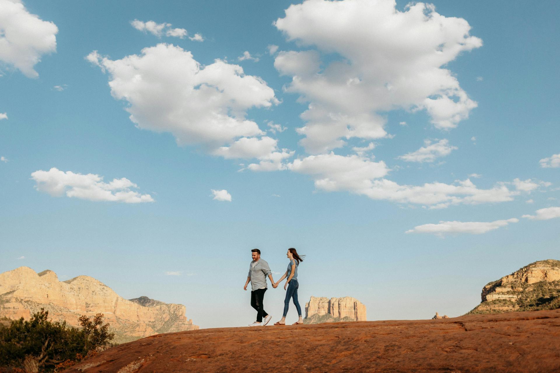 A Man and Woman  Holding Hands Standing while Walking on Brown Rock Formation Under Blue Sky and White Clouds in Sedona Arizona