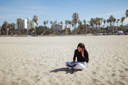 Woman on a sandy beach using a cellphone and handling paperwork, epitomizing remote work and leisure.