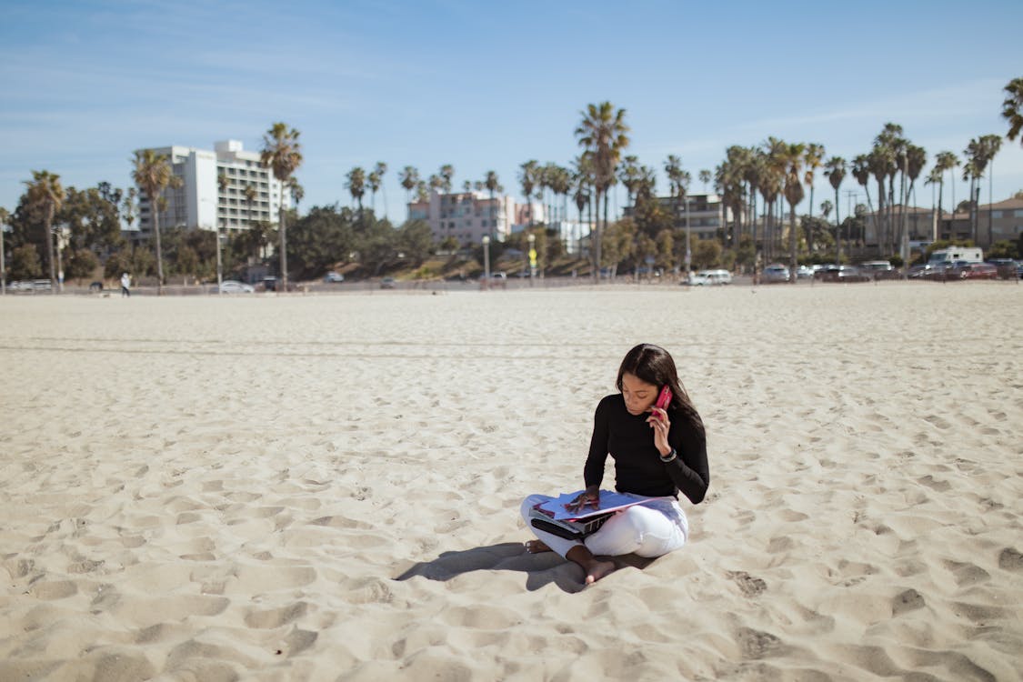 Free Woman on Her Phone Sitting on the Sand Stock Photo