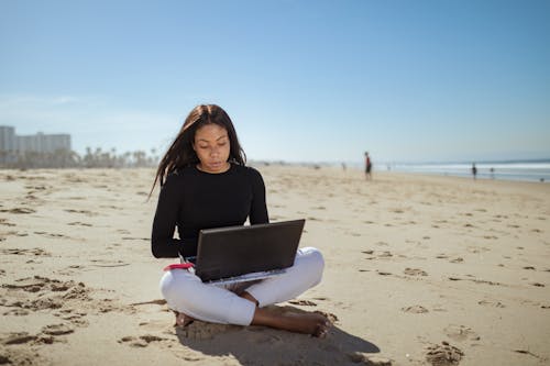 A Woman in Black Long Sleeve Shirt Sitting on the Sand while Using Laptop
