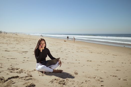 Businesswoman enjoying remote work on a sunny beach using her laptop and smartphone.
