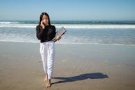 Woman in Black Long Sleeve Shirt and White Pants Holding a Laptop at the Beach