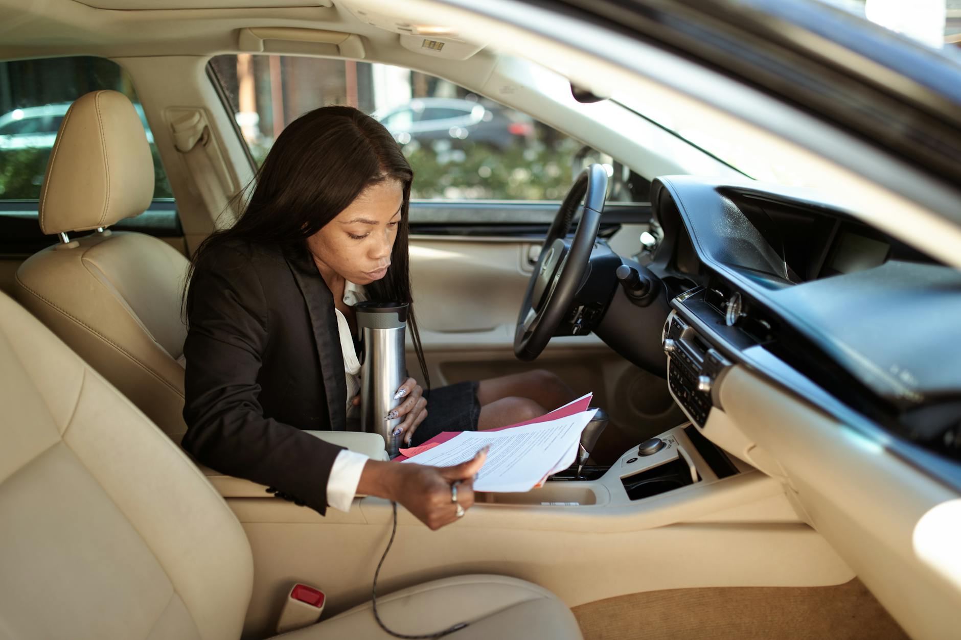 A focused businesswoman reads documents in her car, multitasking with a tumbler.
