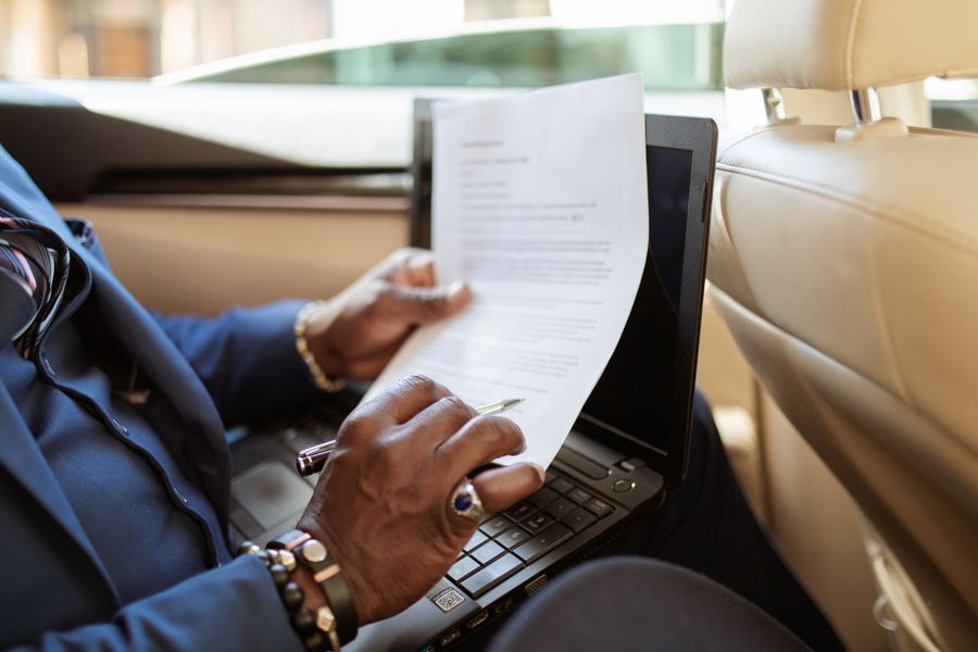 A businessman reviews documents on a laptop while traveling in the backseat of a car.