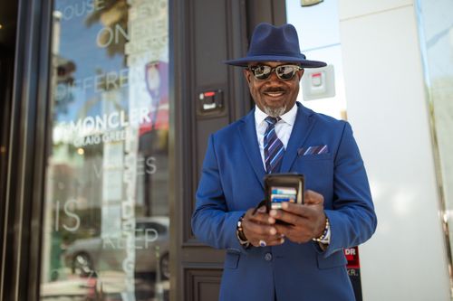 Free A fashionable man in a blue suit and hat smiles while checking his smartphone outside a store. Stock Photo