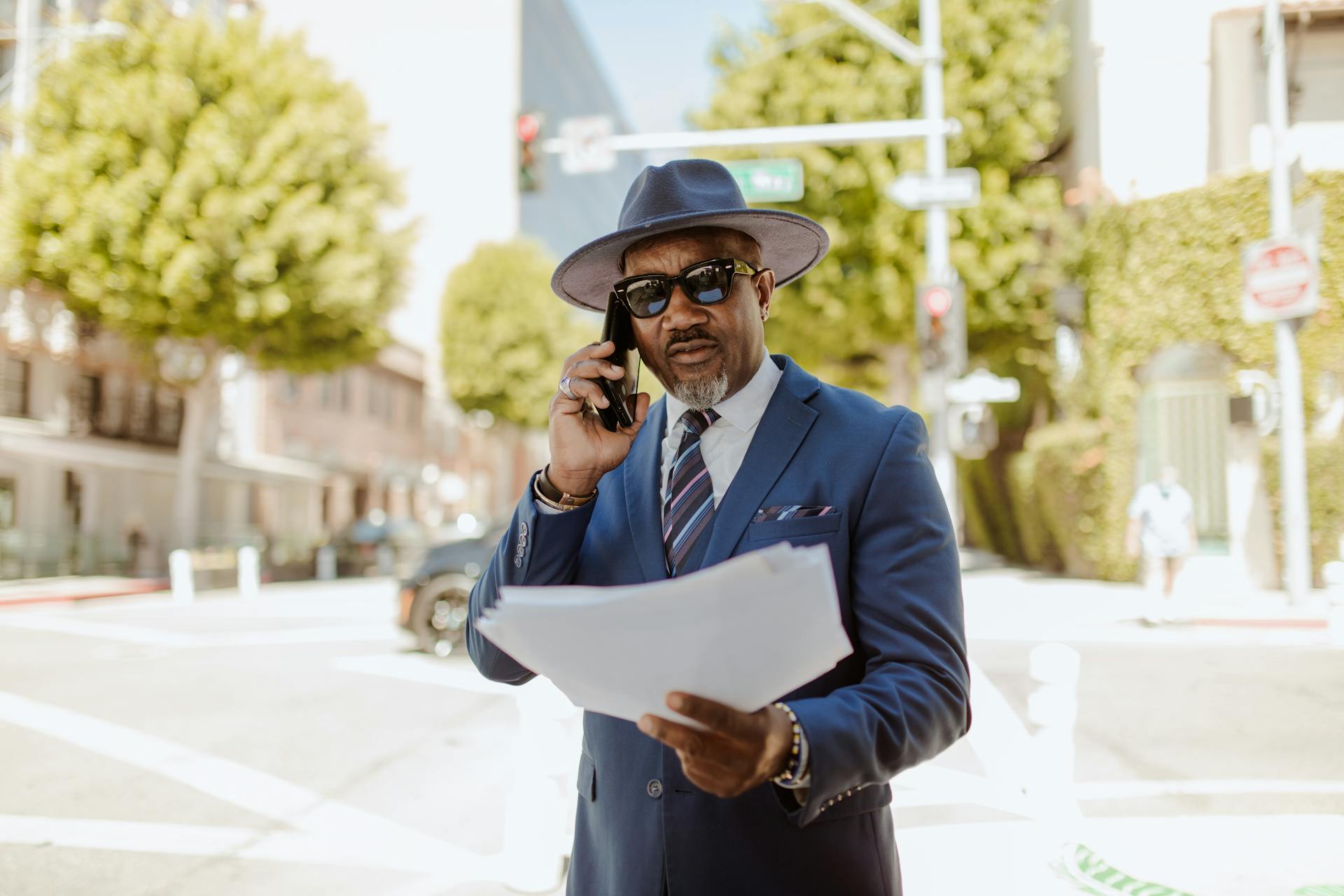 A confident businessman in a blue suit and hat talks on the phone while holding documents outdoors.