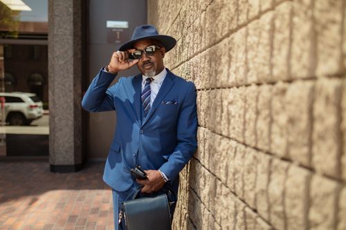 Free Confident businessman in a blue suit and hat posing against a modern building wall. Stock Photo