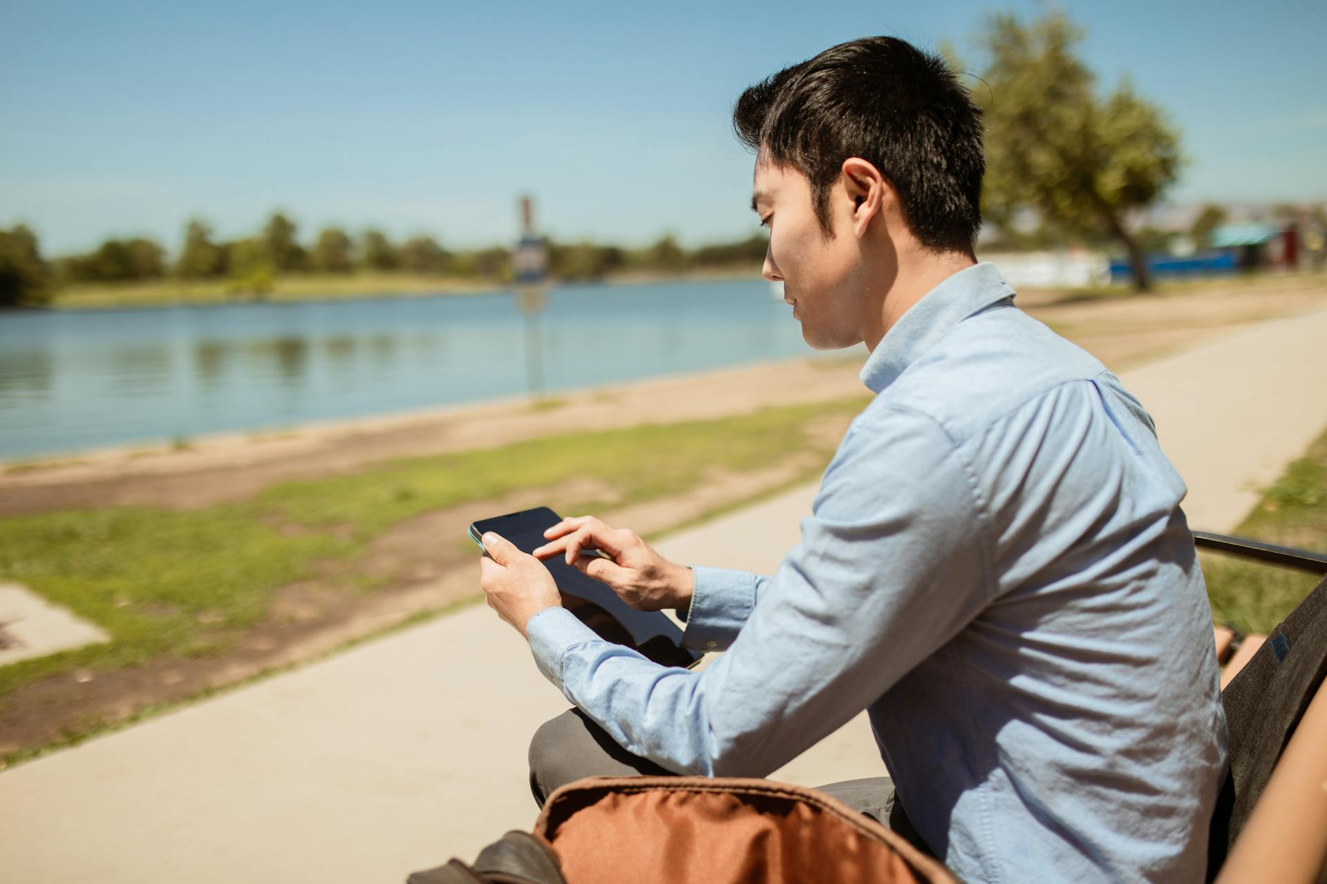 Asian man in blue shirt using a digital tablet while sitting on a riverside bench outdoors.