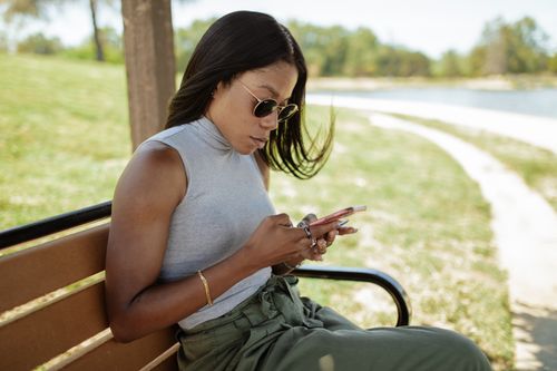 Free A fashionable woman with sunglasses uses her smartphone on a sunny park bench. Stock Photo