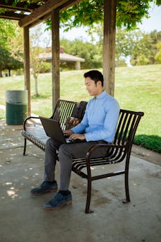 Asian man in business attire working on laptop in a sunny park. Ideal for remote work themes.