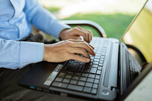 Hands typing on a laptop outdoors, showcasing remote work or study in a sunny environment.