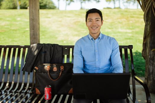 Asian man in casual attire working on laptop outside on a sunny day, enjoying remote work lifestyle.