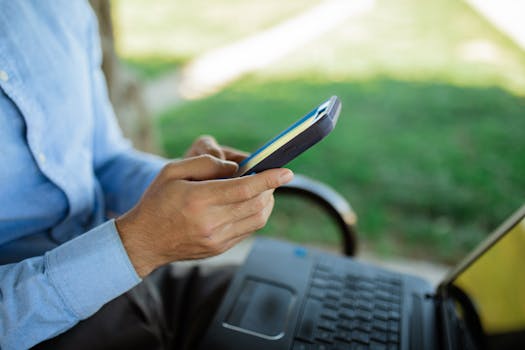 A person uses a smartphone while seated outdoors with a laptop, showcasing remote work lifestyle.