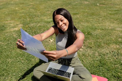 Free Joyful woman sitting on grass with laptop and paper, enjoying outdoor work. Stock Photo