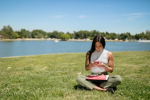 Free A woman sits on grass in a park by the lake, using her smartphone on a sunny day. Stock Photo
