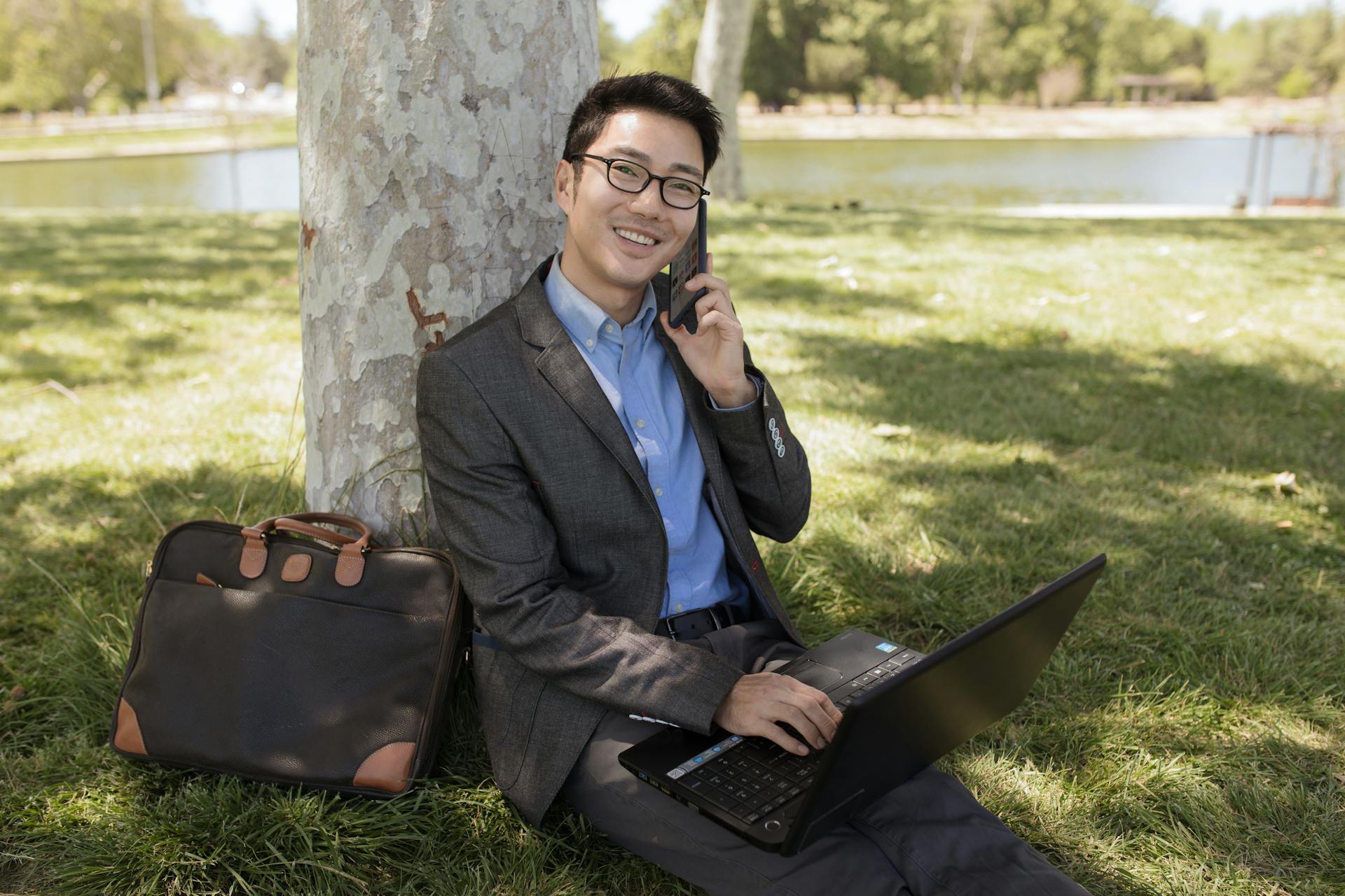 Smiling businessman in corporate attire using laptop and phone under a tree in a park.