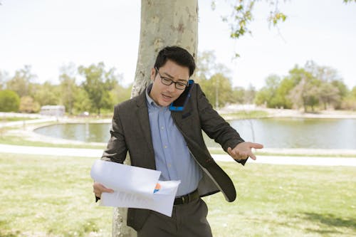 A Man Talking on a Smartphone while Looking at Documents in a Park