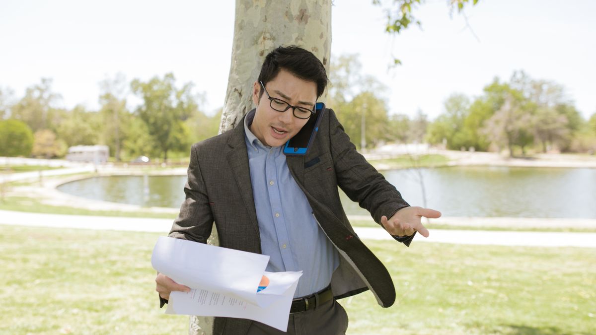 Asian businessman outdoors multitasking with phone and documents in park setting.