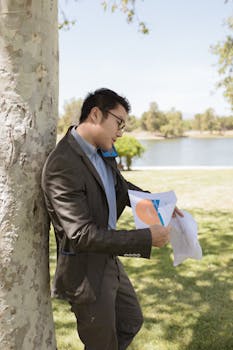 Professional in corporate attire holding documents, speaking on phone outdoors by lake.