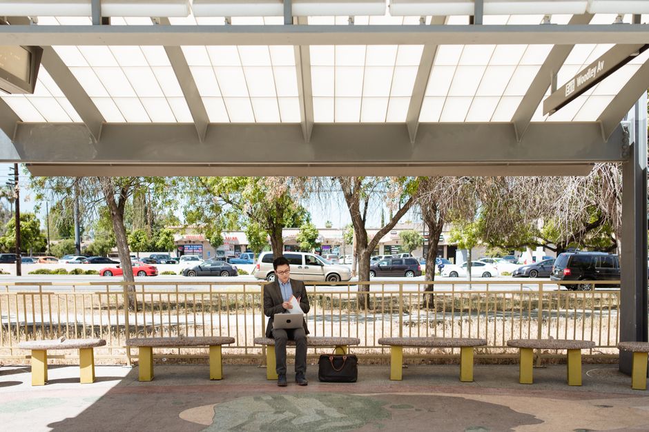 A businessman in corporate attire working on a laptop at a bus stop on a sunny day.