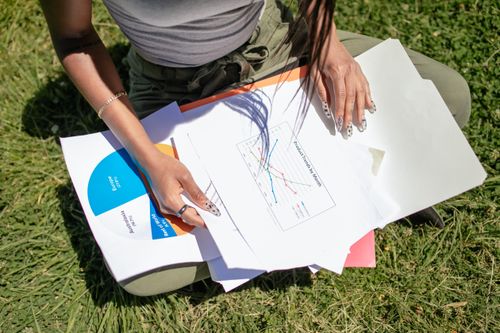Free A woman sits on grass reviewing charts and business documents in a park setting, capturing an outdoor work scene. Stock Photo