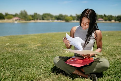 Free A woman sits on grass by a lake, examining papers on a sunny day. Stock Photo