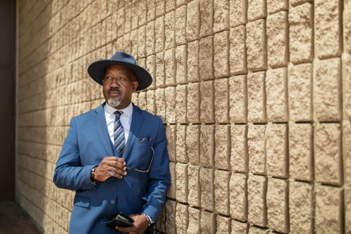 Free Confident businessman in blue suit and hat leaning against textured wall outdoors. Stock Photo