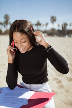 Woman smiling while talking on phone at a sunny beach with documents.