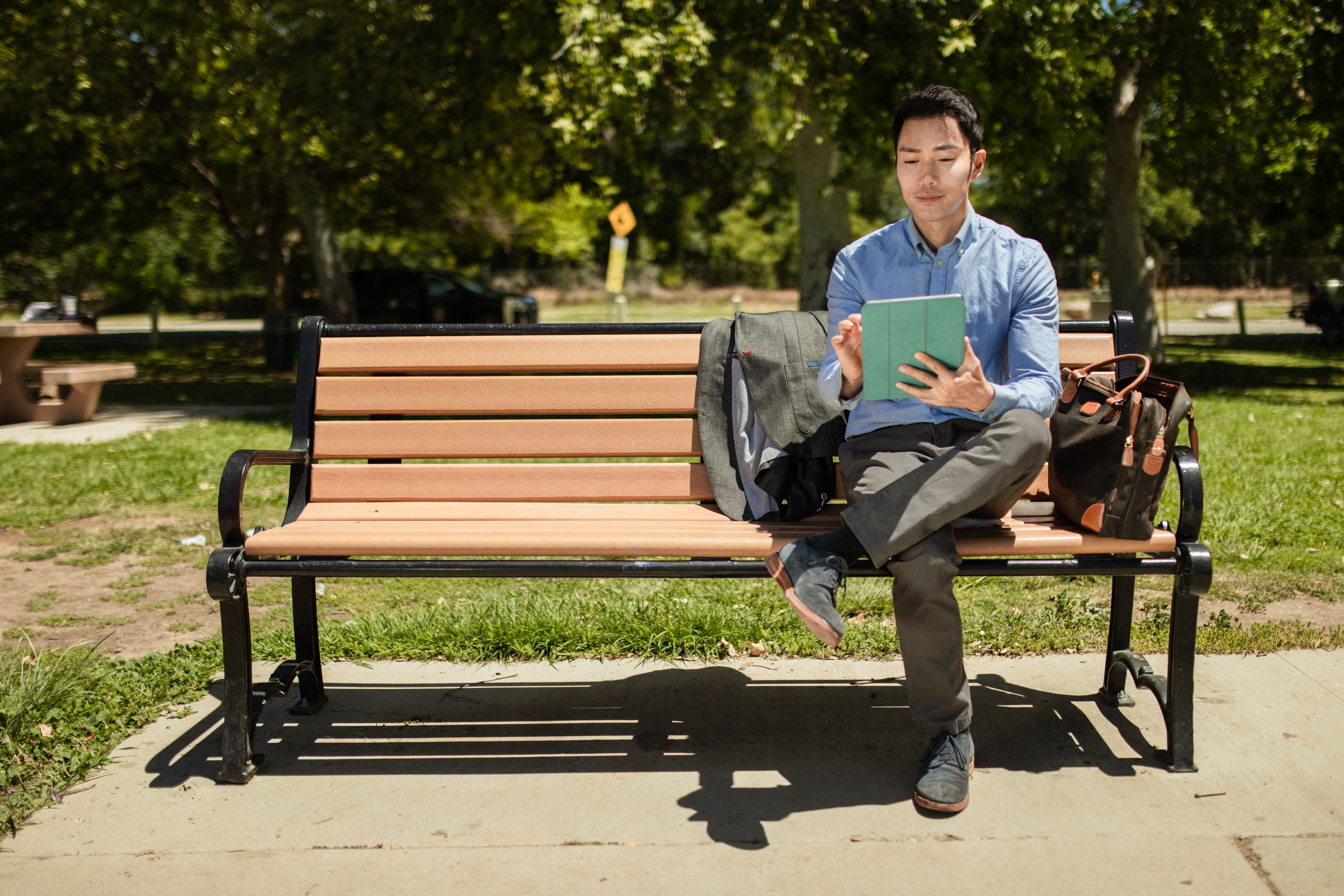 An adult man sits on a bench in a park, using a tablet on a sunny day.