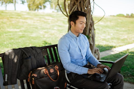 Asian businessman sitting on a bench typing on a laptop, focused on remote work in a sunny park setting.
