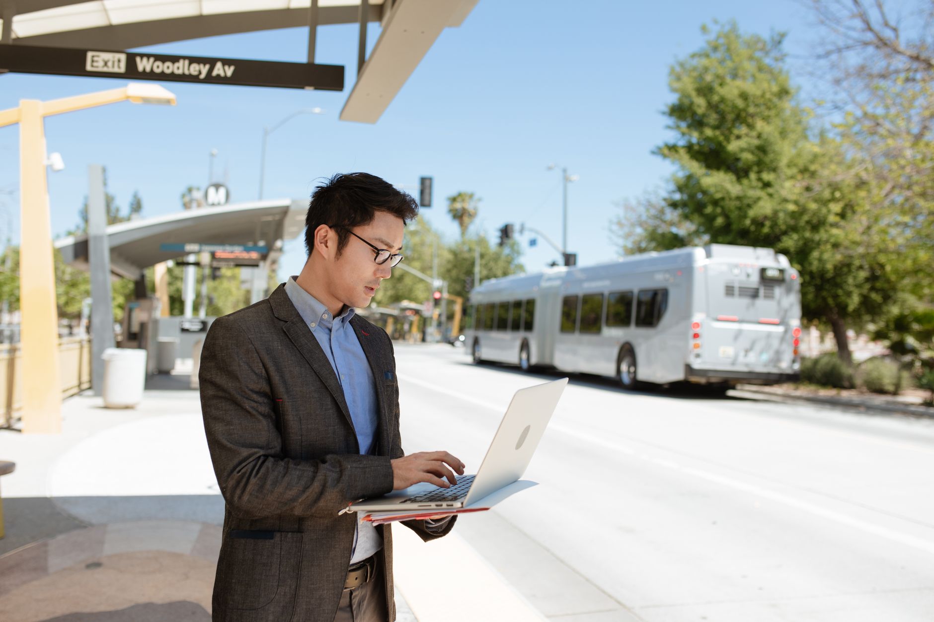 Asian businessman in a black suit working on a laptop at a bus stop on Woodley Avenue.