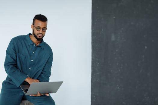 Young man with beard in blue sits typing on a laptop indoors.