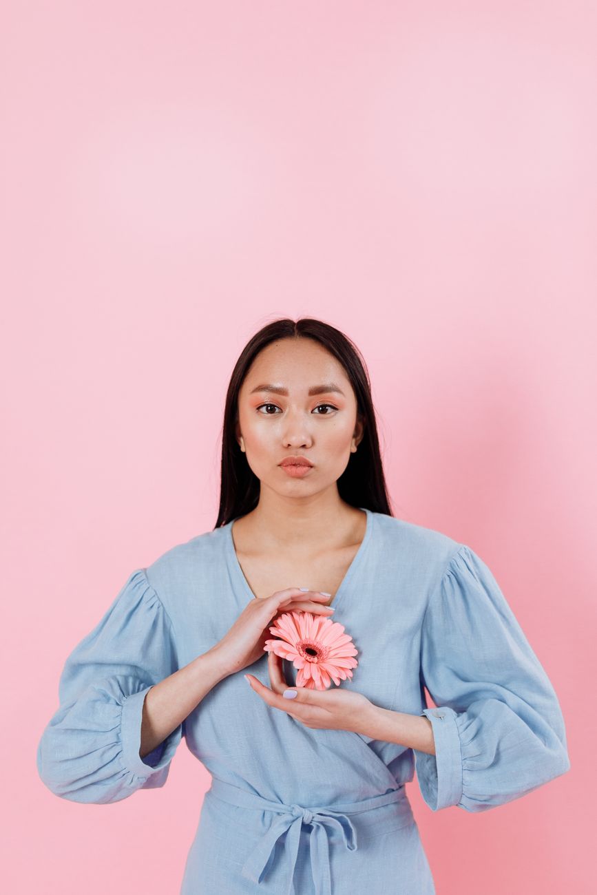 Fashionable young Asian woman in blue outfit with pink flower poses against light pink background.