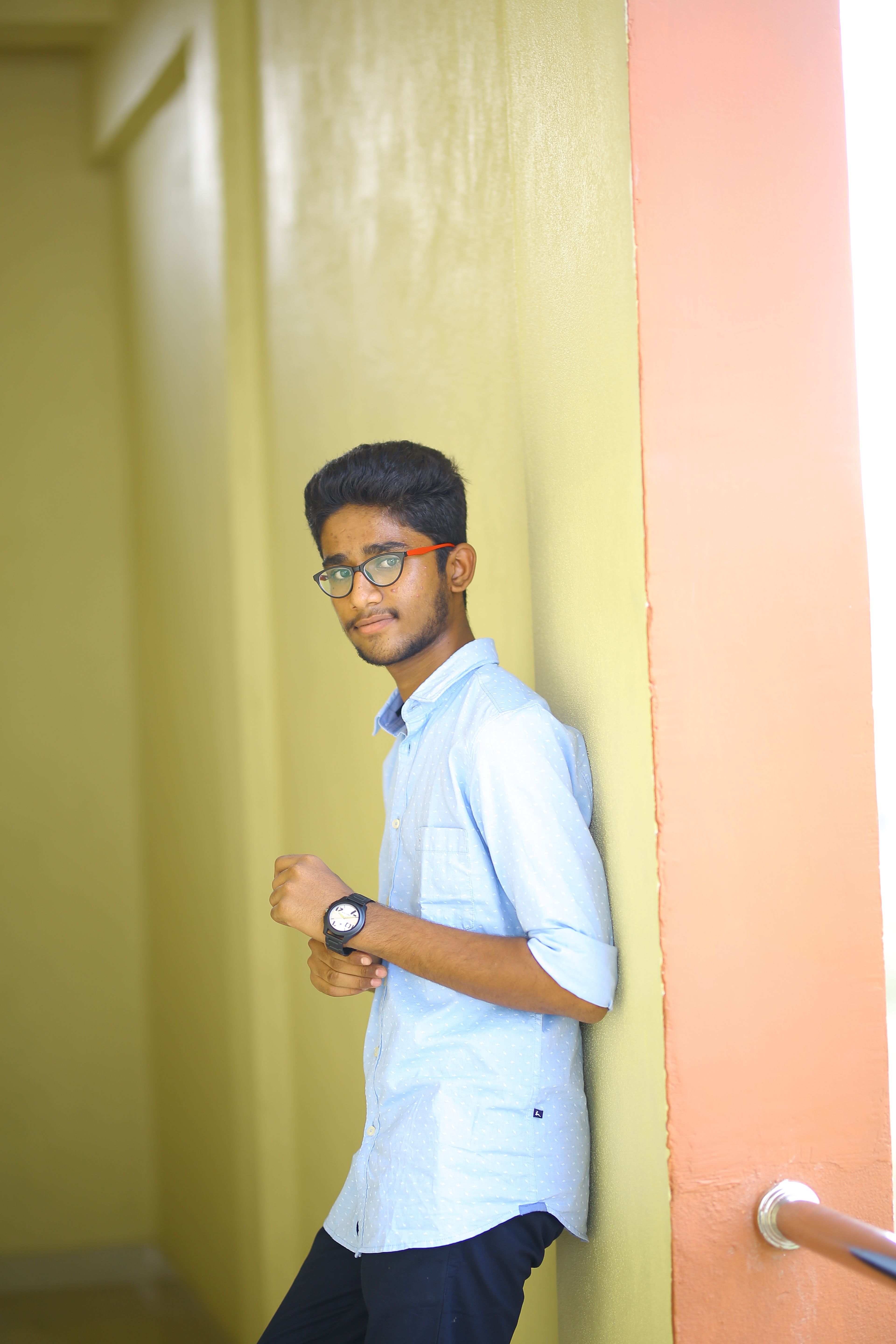 Portrait of a young man with glasses indoors, leaning against a wall with soft lighting.