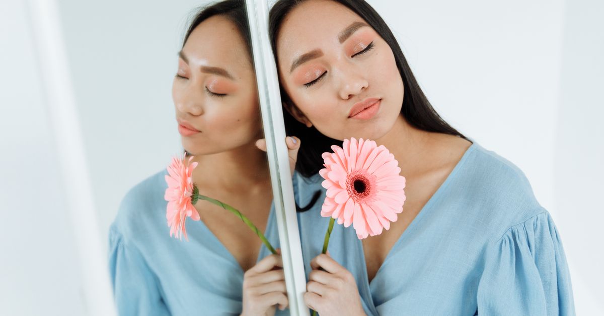 Portrait of a woman with eyes closed, holding a pink flower and its reflection in a mirror.