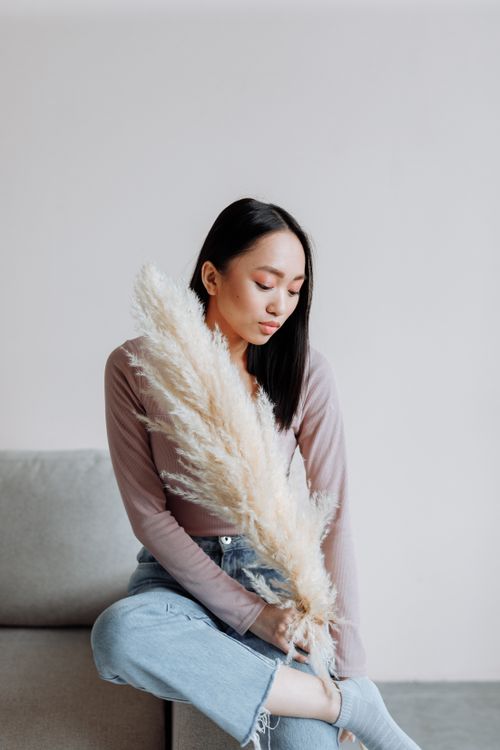 Free A stylish adult woman sits in a minimalist room holding pampas grass, showcasing modern casual fashion. Stock Photo
