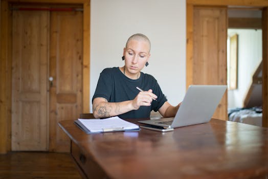 Bald woman with tattoos working from home using a laptop and taking notes. Indoor setting.