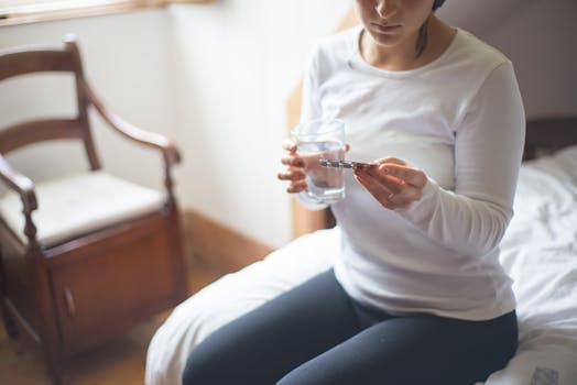 A woman in a white shirt sitting on a bed holds a glass of water and medicine.