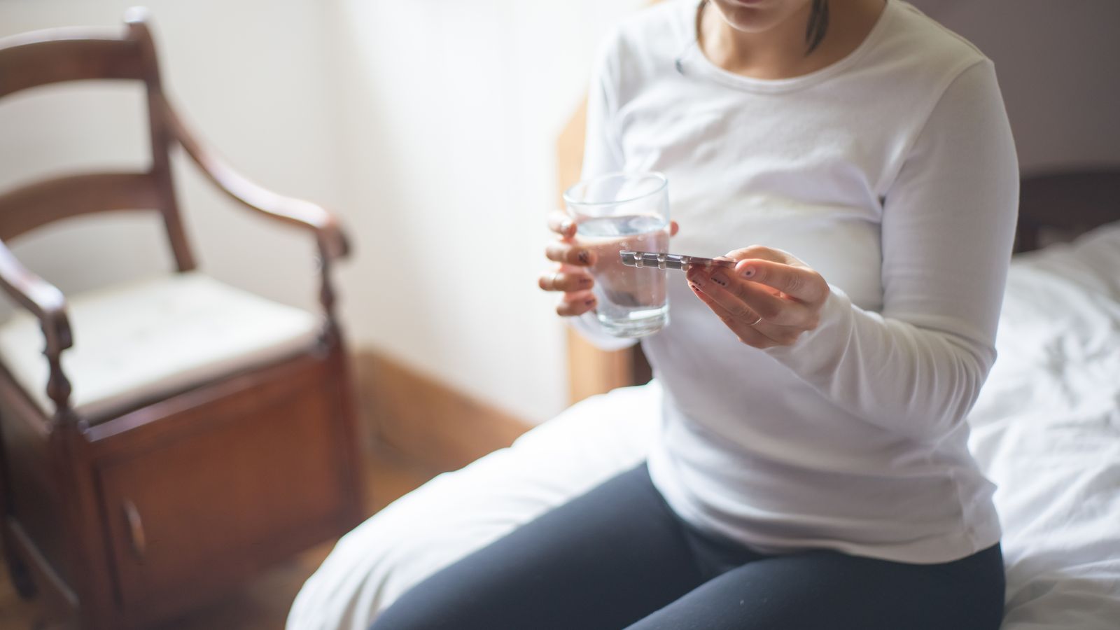 A woman in a white shirt sitting on a bed holds a glass of water and medicine.