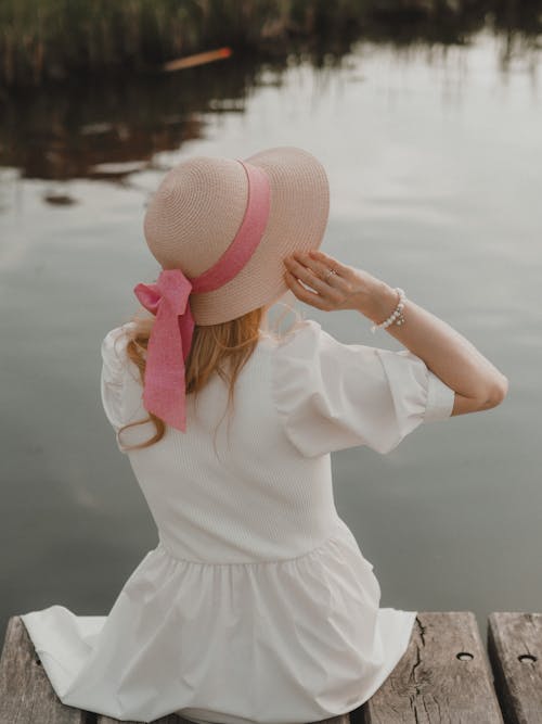 Back view of unrecognizable female in white dress sitting on wooden pier and adjusting hat near calm lake in daytime