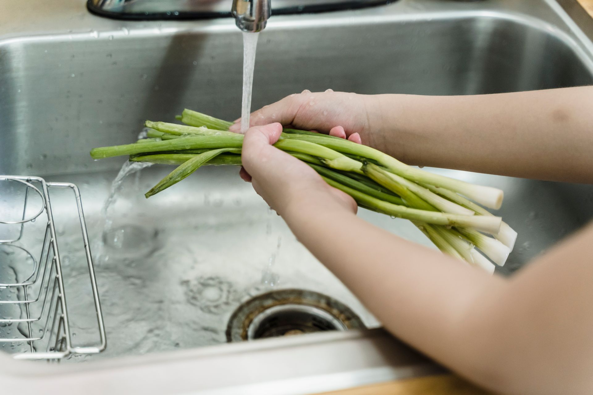 Hands washing fresh leeks under running water in a kitchen sink, ensuring hygiene and cleanliness.