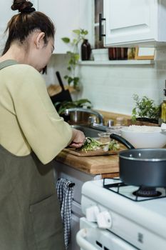 Asian woman chopping vegetables while cooking in a cozy kitchen setting, highlighting food preparation.