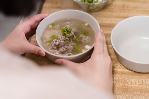 Close-up of a bowl of pork soup with green onions held indoors, highlighting warmth and comfort.