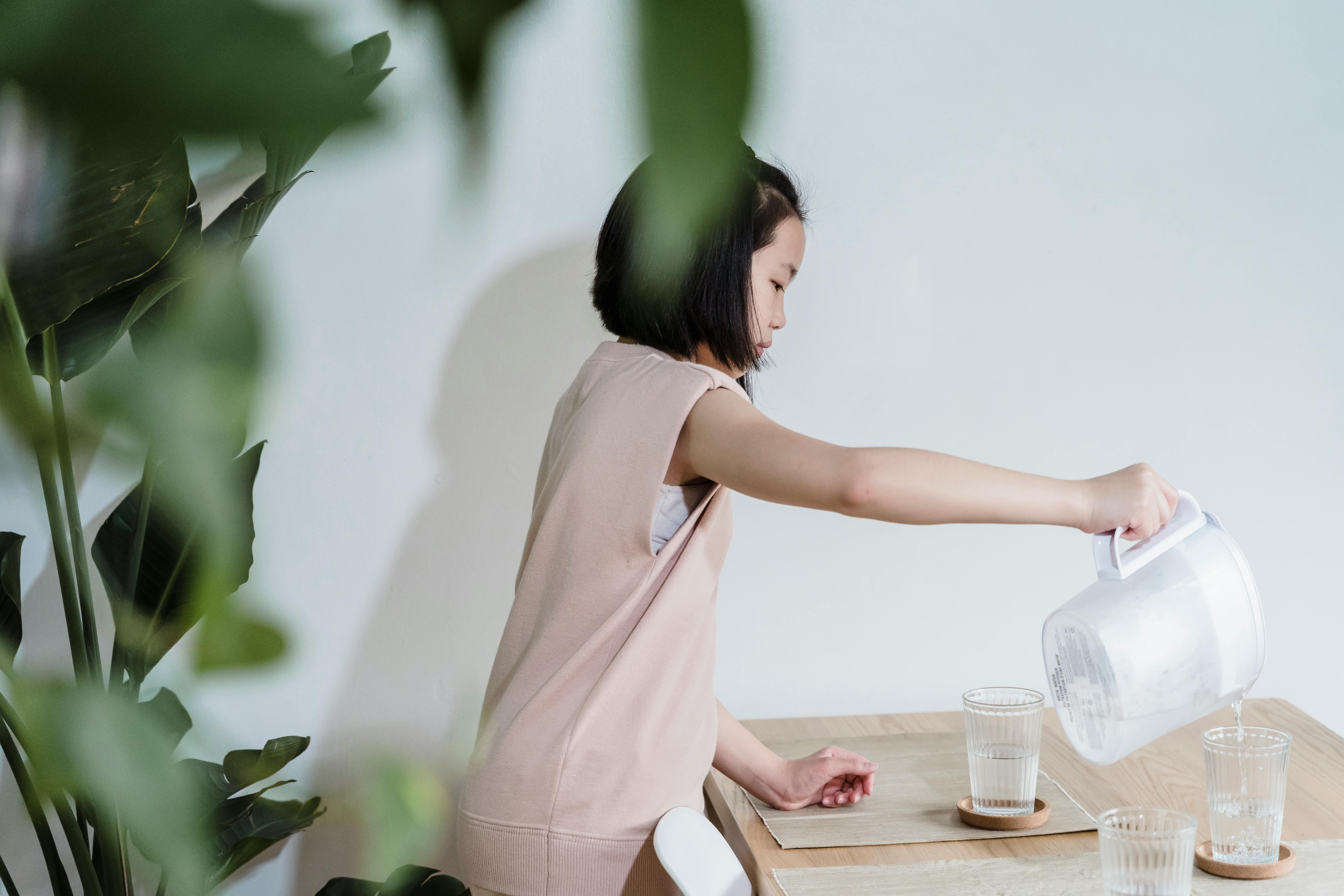 A young girl pours water from a pitcher into glasses at a wooden table indoors.